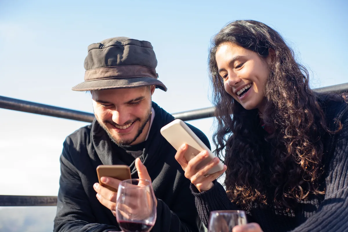 Dos clientes jóvenes sonriendo mientras reservan restaurante desde el móvil por WhatsApp en una terraza de verano